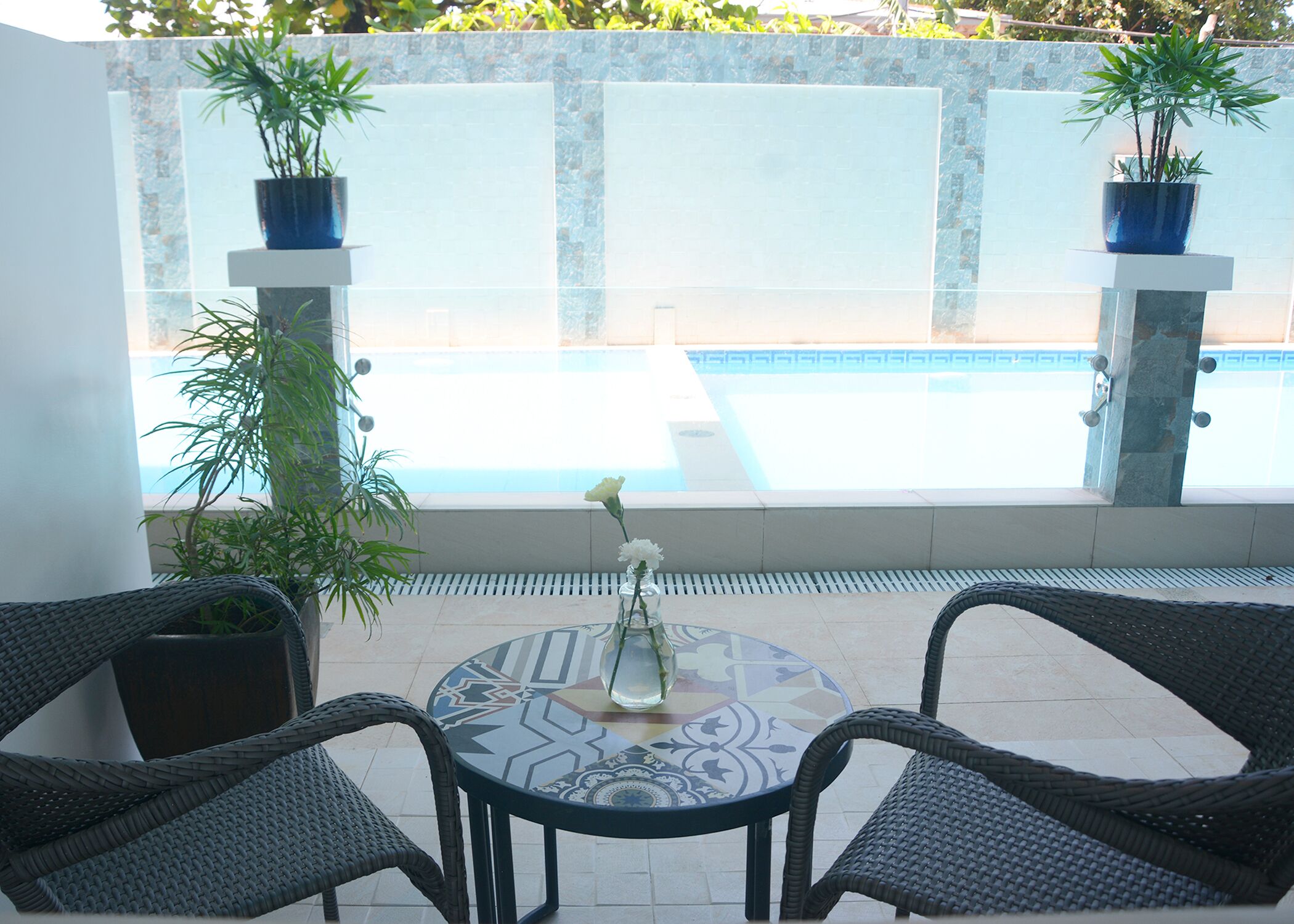 Un patio con sillas de mimbre, una mesa con mantel estampado y un jarrón con flores. Hay dos plantas en macetas sobre pedestales y se ve una piscina al fondo.