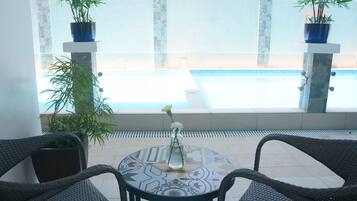 Un patio con sillas de mimbre, una mesa con mantel estampado y un jarrón con flores. Hay dos plantas en macetas sobre pedestales y se ve una piscina al fondo.