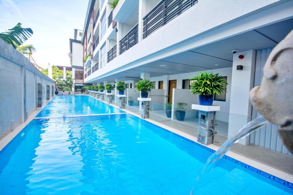 Una piscina con superficie de agua azul cristalina, rodeada por un edificio moderno con balcones y plantas en macetas.