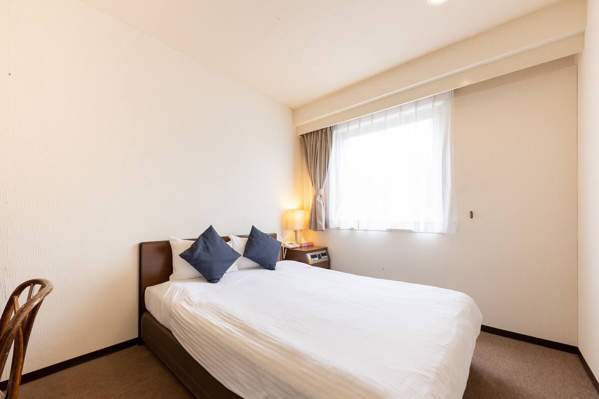 A hotel room with a bed, two pillows, a nightstand, a lamp, and a window with curtains.