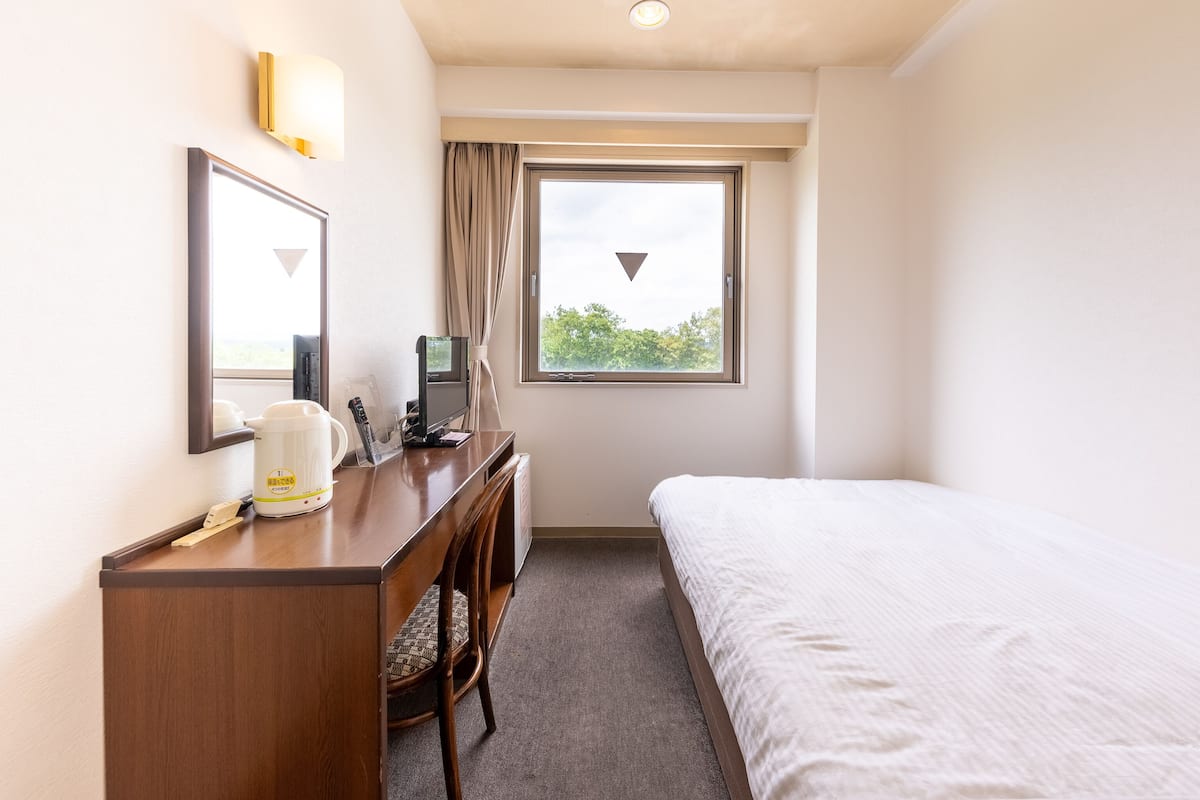 A hotel room with a bed, a desk with a computer, a coffee maker, and a window with a view of trees.