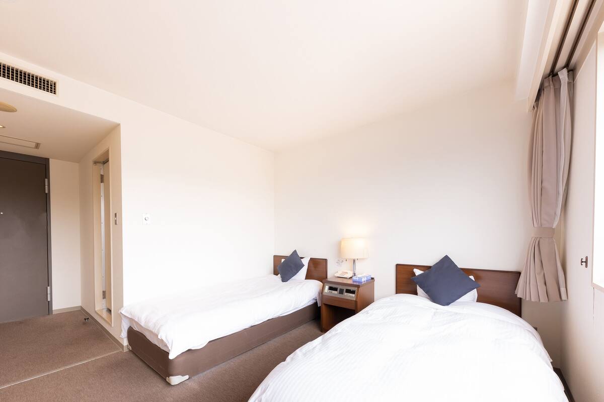 A hotel room with two beds, a bedside table with a lamp, and a door.