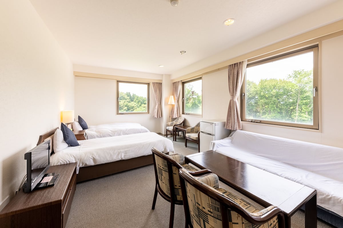 A hotel room with two beds, a desk, a chair, a small table, and large windows with curtains.
