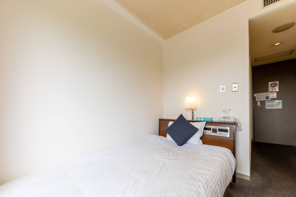 A hotel room with a bed, a nightstand with a lamp, a telephone, and a door with signs.