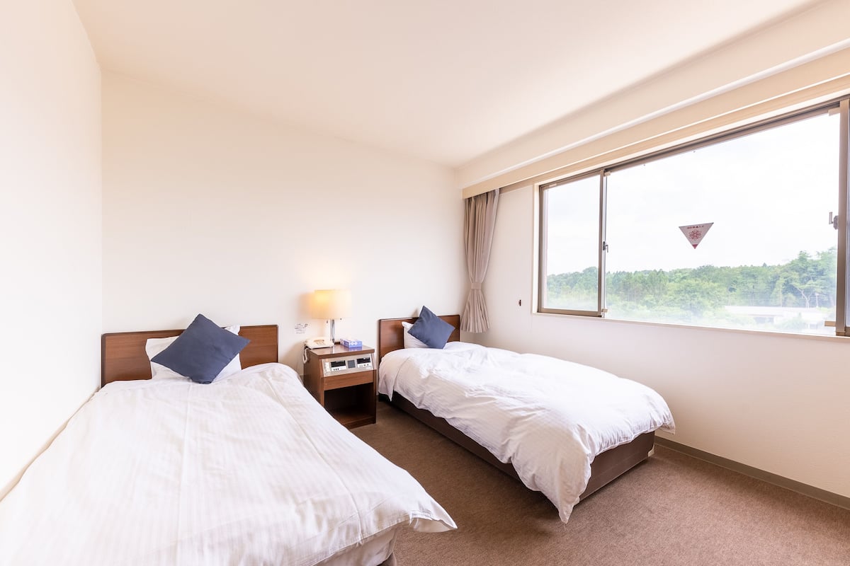 A hotel room with two single beds, a nightstand with a lamp, and a large window offering a view of greenery.