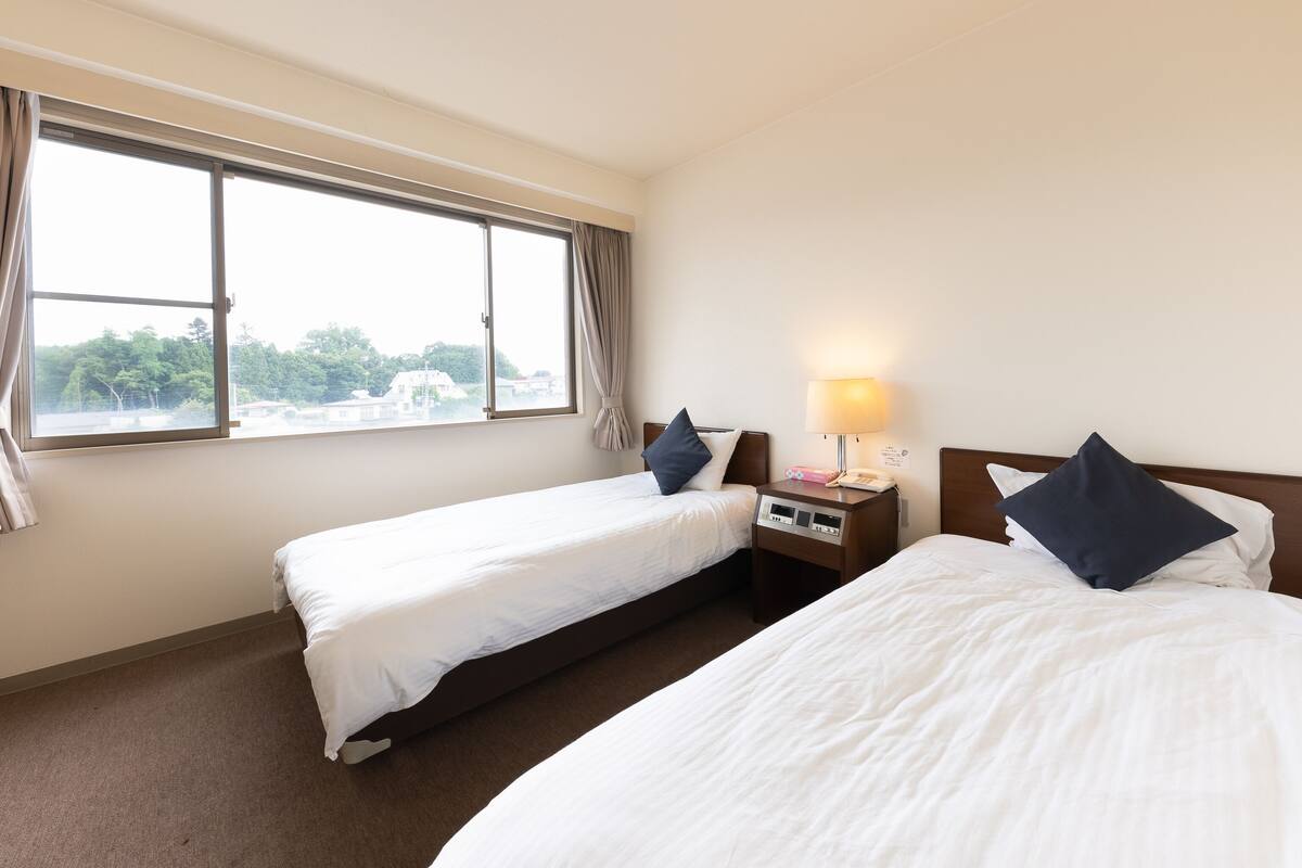A hotel room with two beds, a nightstand with a lamp, and a window with curtains.