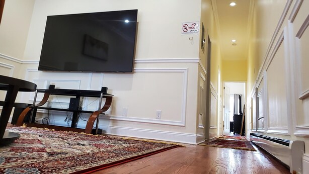 A spacious hallway with a flat-screen TV mounted on the wall, a dining area with a table and chairs, and a large patterned rug on the floor.