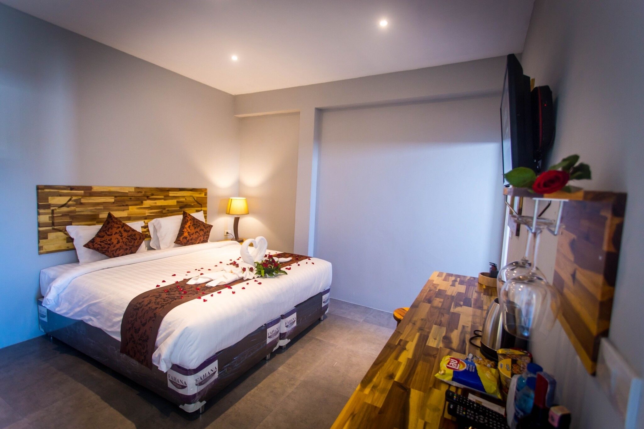 A hotel room with a neatly made bed, a wooden headboard, a bedside lamp, and a wooden desk with a glass vase.