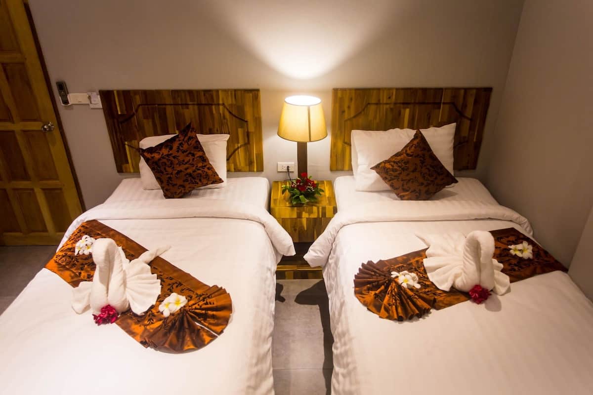 A hotel room with two beds, each with a wooden headboard, white bedding, and decorative pillows. A bedside table with a lamp and a flower arrangement is between the beds.