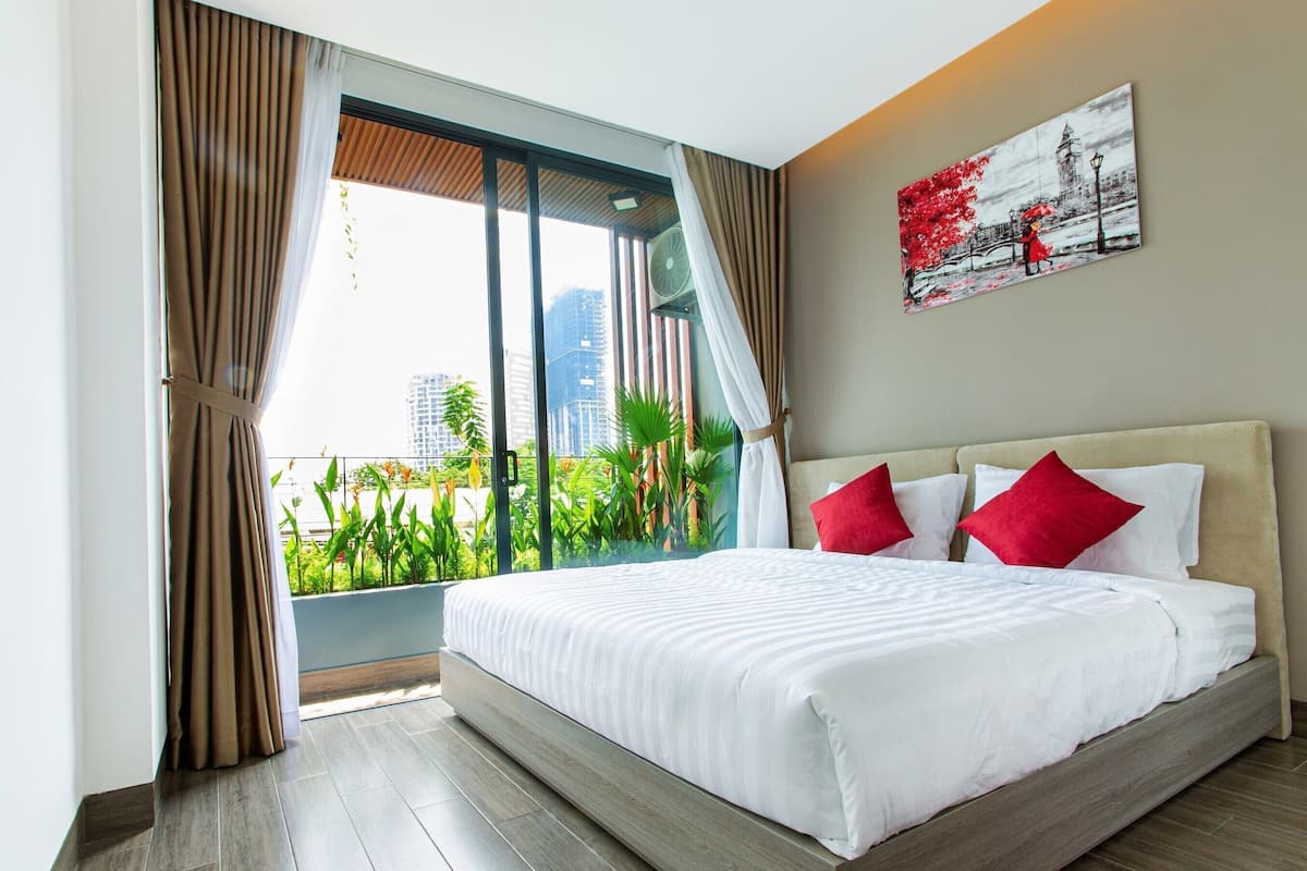 Une chambre d’hôtel avec un grand lit, une vue sur la verdure et les bâtiments, et un tableau accroché au mur.