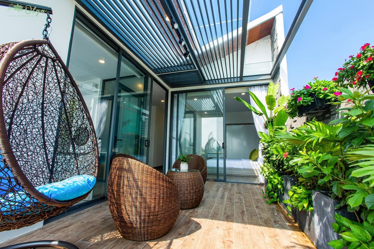Un espace extérieur moderne, aménagé avec des meubles en osier et un fauteuil suspendu, entouré d’une végétation luxuriante et doté d’une entrée en verre.