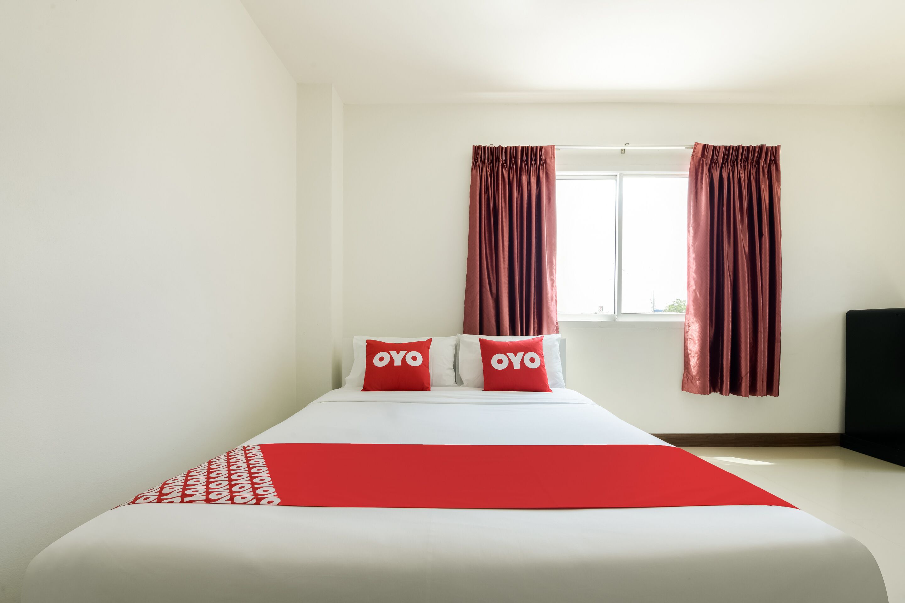 Habitación de hotel con cama, dos almohadas con la marca 'OYO', una colcha roja y una ventana con cortinas.