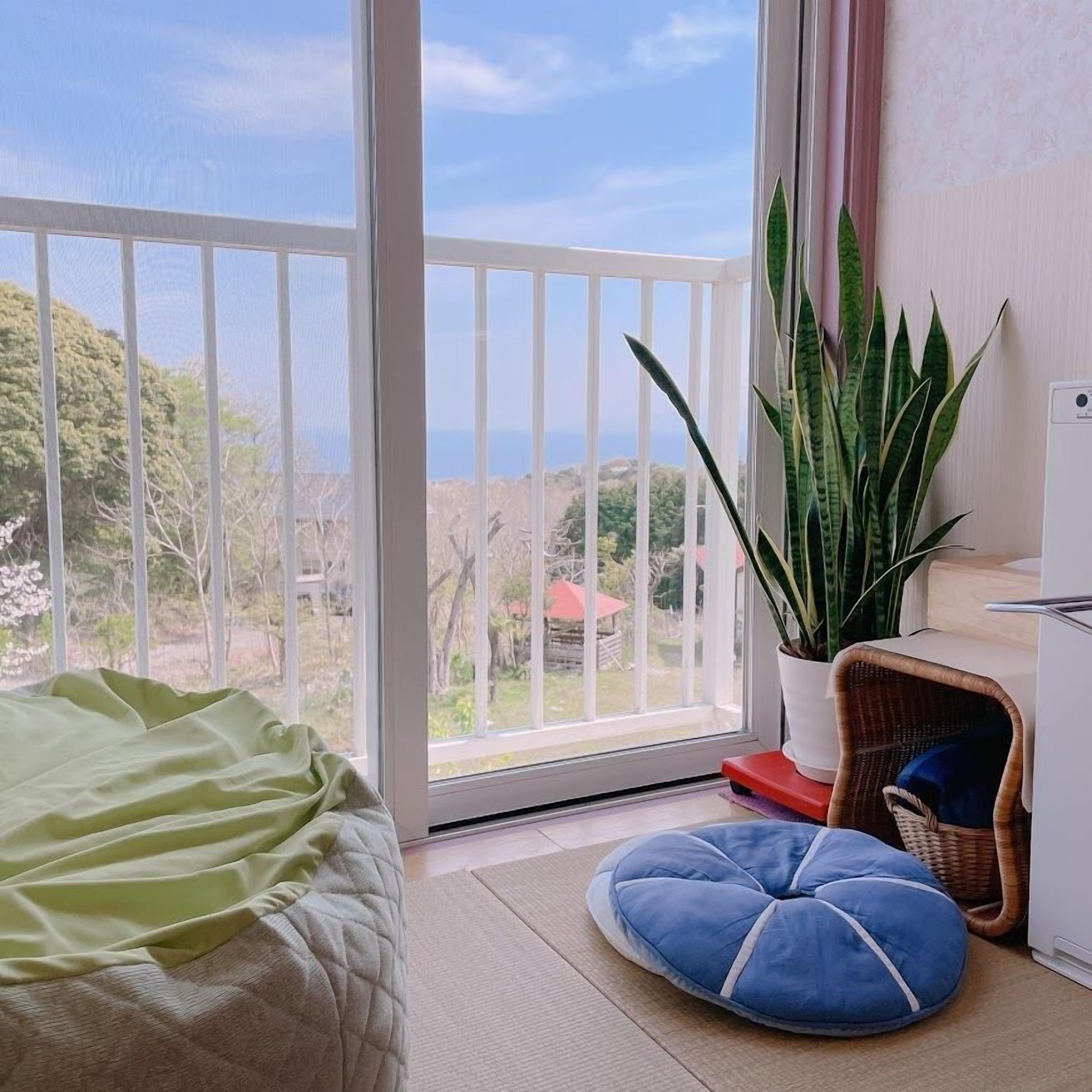 Ein Balkon mit Blick auf die Landschaft, ein Bett, ein Blumentopf, ein Korbstuhl und ein weißes Schränkchen.