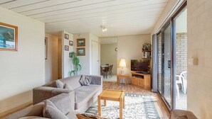 A living room with a sofa, a coffee table, and a TV on a stand. There is a dining area with chairs and a table in the background. - Apollo Unit 27 - Ground Floor (Narooma)