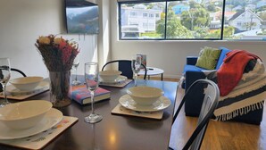 A dining area with a table set for two, a vase with flowers, and a view of houses through a large window. - U Suites Kilbirnie (Wellington)