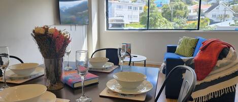 Un comedor con una mesa dispuesta para dos, un jarrón con flores y vistas a casas a través de un gran ventanal.