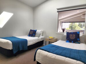 A bedroom with two beds, each with a blue blanket and a pillow with a 'U' design. There is a window with a view of buildings and trees, and a small table with a book and a cup. - U Suites Kilbirnie (Wellington)