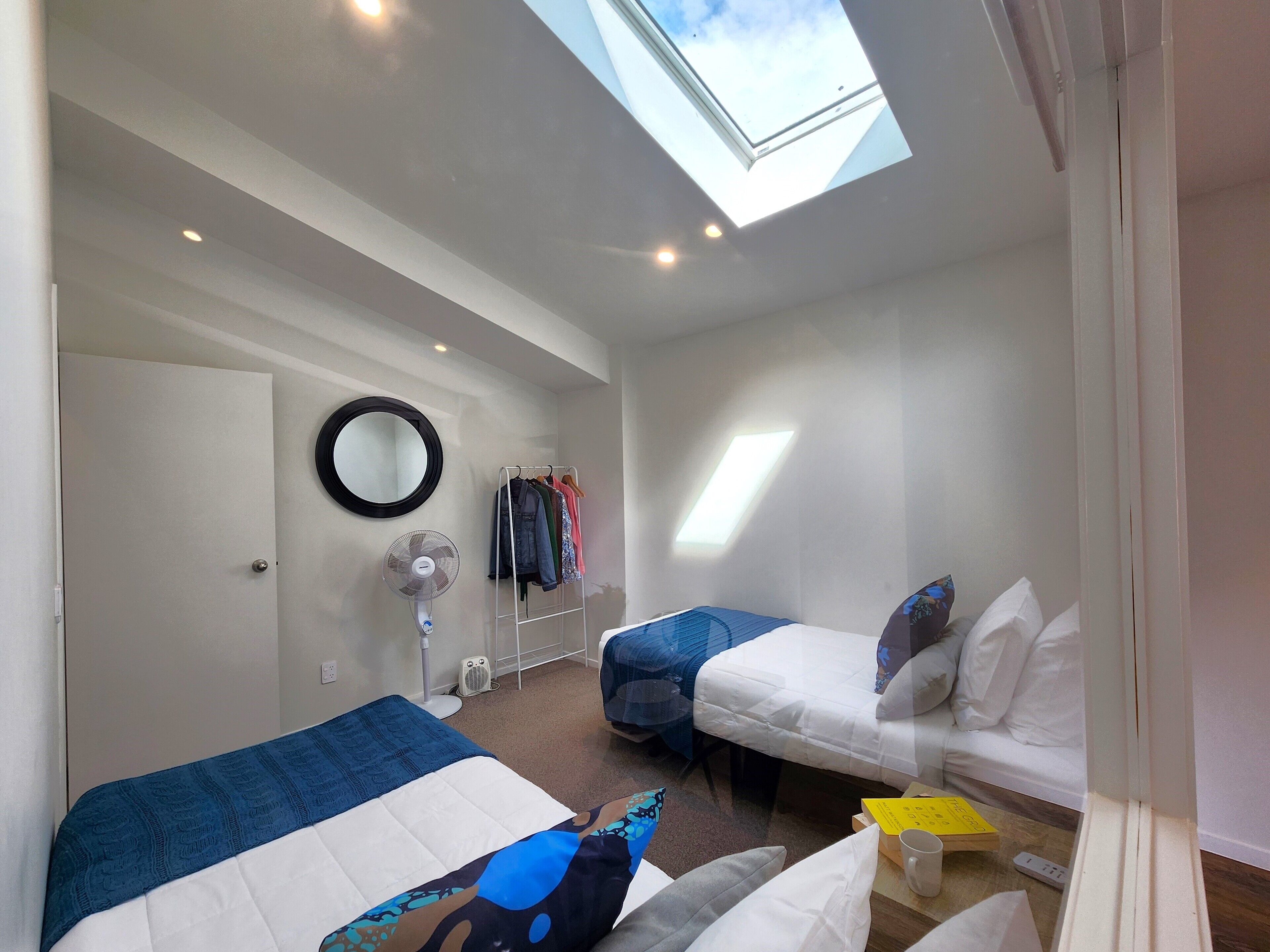 Un dormitorio pequeño con dos camas, un tragaluz, un espejo, un ventilador y una percha para ropa.