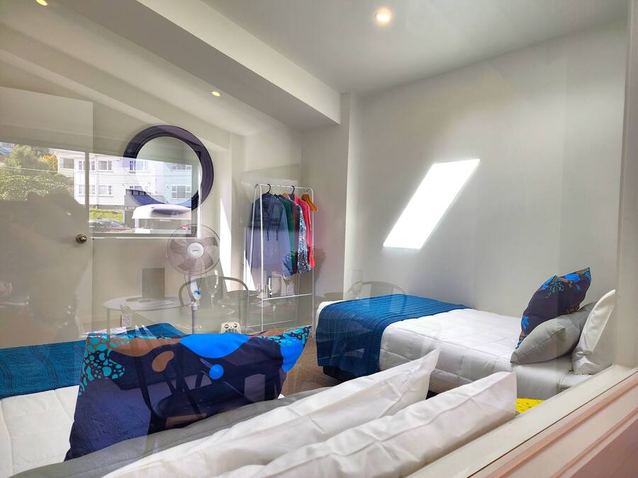 A modern hotel room with a glass partition, a bed with white bedding, a blue blanket, and a small table with a fan.