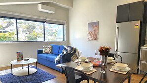 A modern living room with a blue sofa, a dining table set with white plates and glasses, and a kitchen area with a refrigerator. - U Suites Kilbirnie (Wellington)