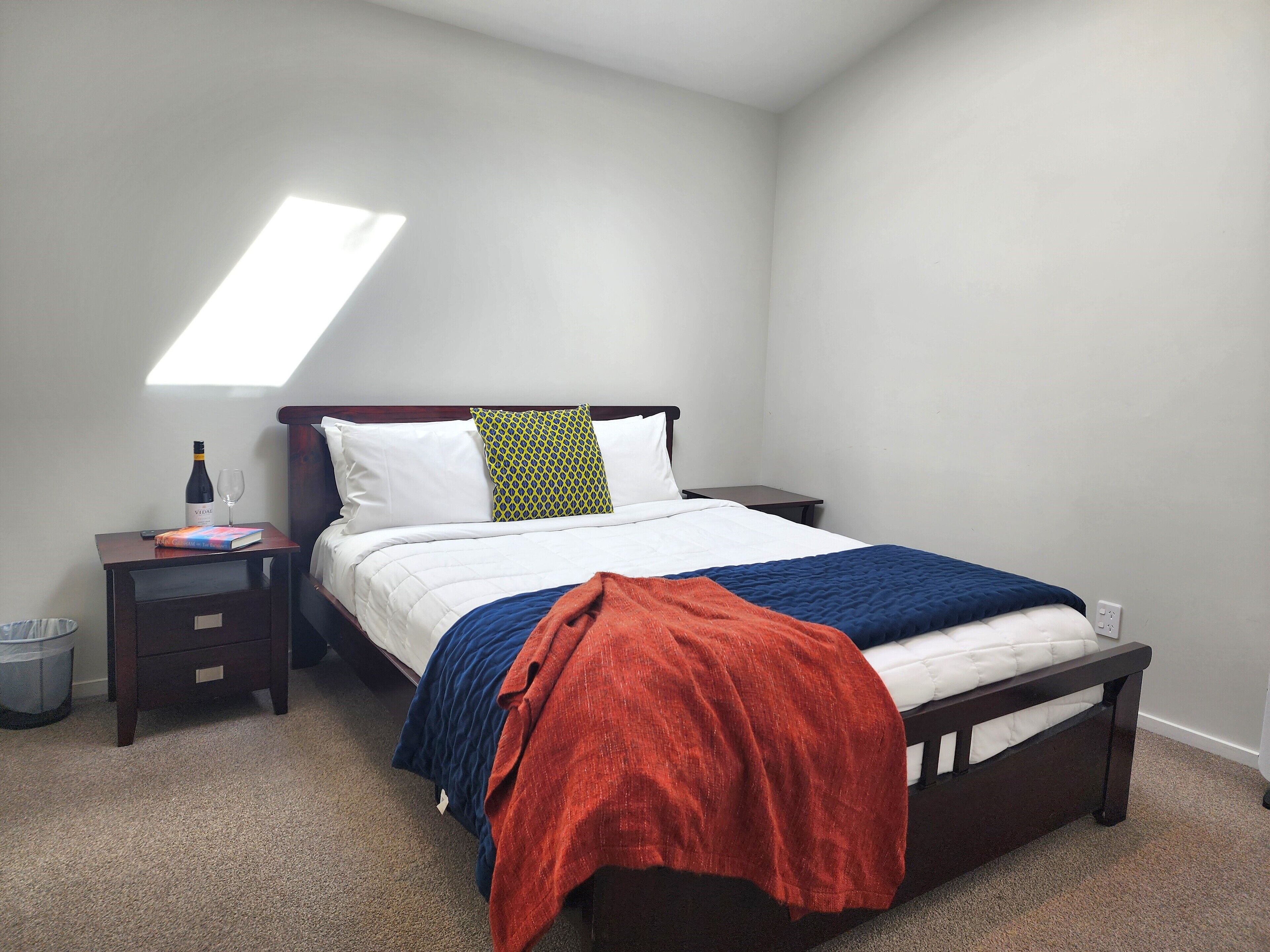 A neatly arranged bedroom with a bed, a bedside table, a nightstand, and a small trash bin.