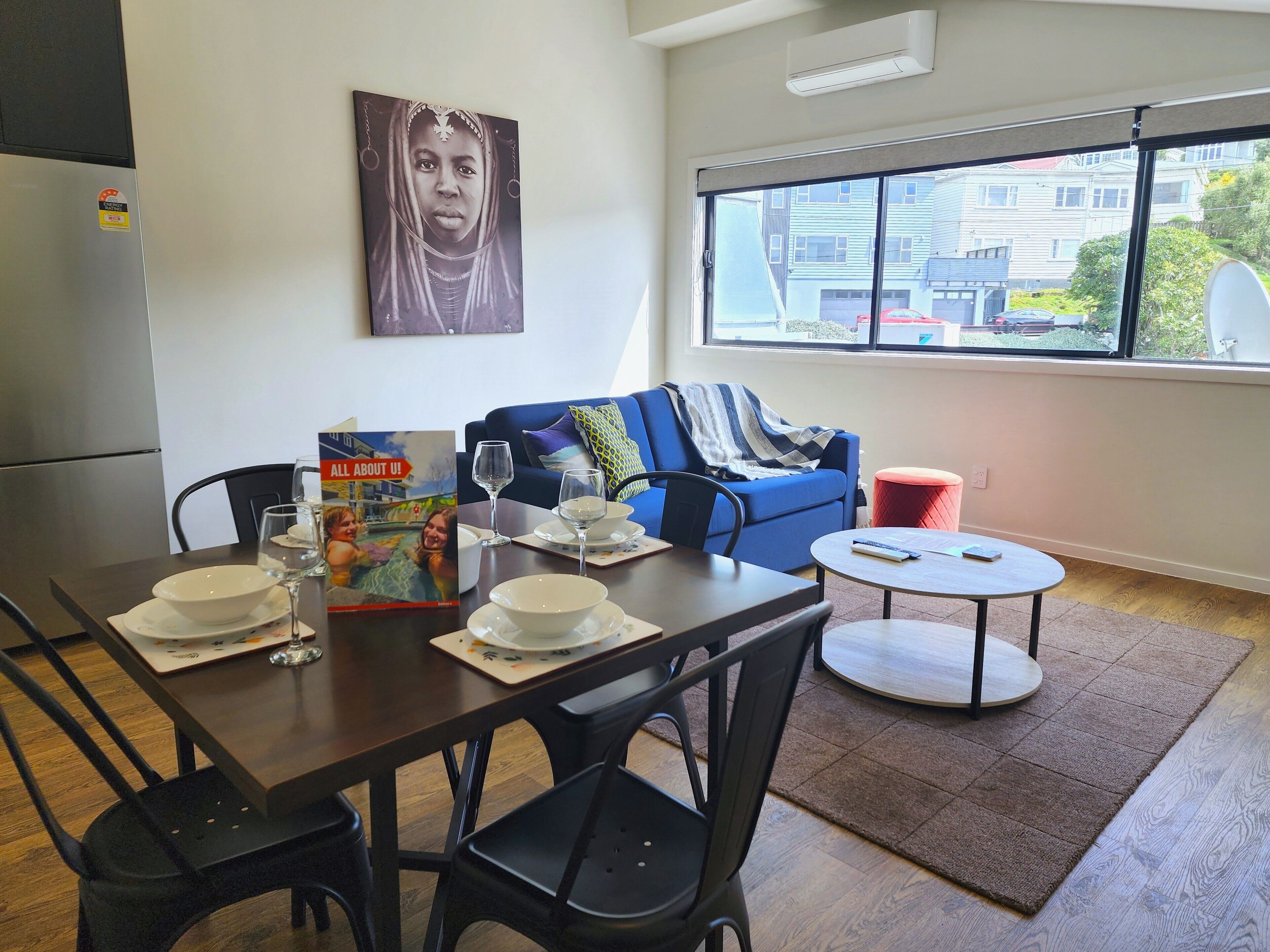 A modern living room with a dining area, featuring a sofa, a red stool, a round table, and a large window with a view of buildings.