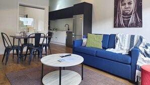 A modern living room with a blue sofa, a round coffee table, and a dining area with a table and chairs. - U Suites Kilbirnie (Wellington)