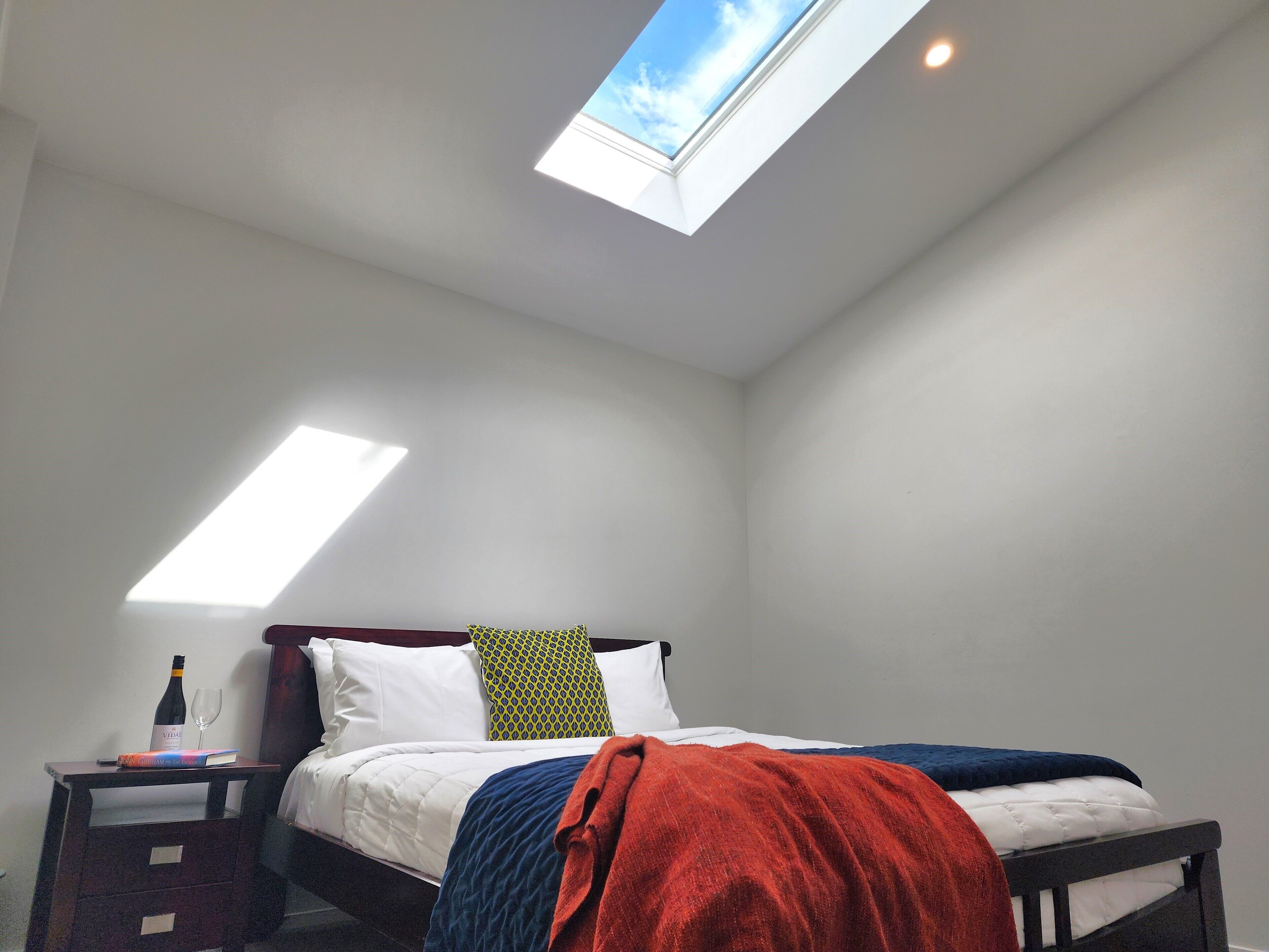 Une chambre avec un puits de lumière, un lit avec une literie blanche, une couverture rouge et une table de chevet sur laquelle se trouvent une bouteille et des verres.