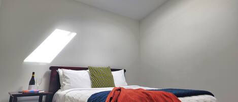 Une chambre avec un puits de lumière, un lit avec une literie blanche, une couverture rouge et une table de chevet sur laquelle se trouvent une bouteille et des verres.