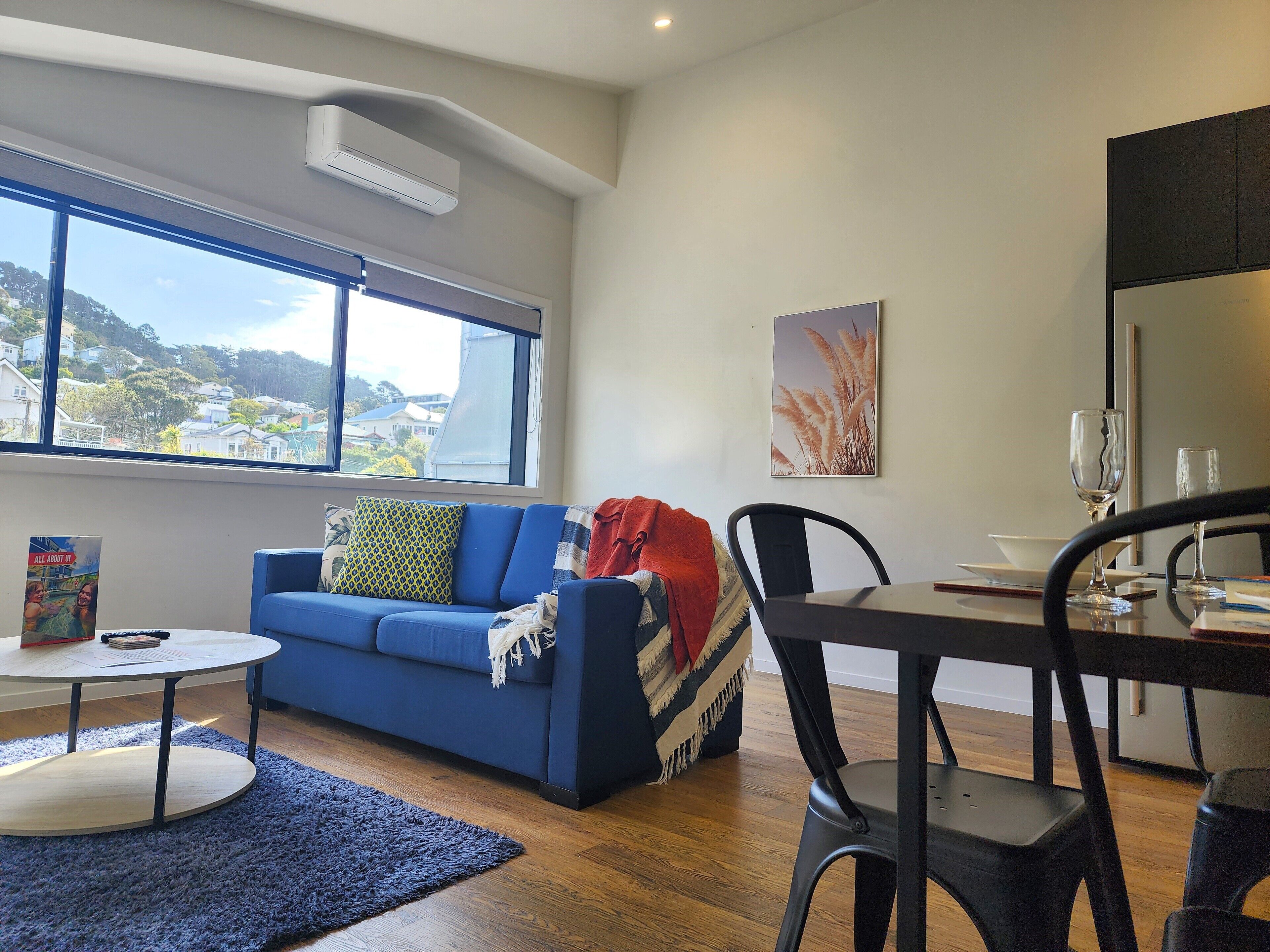 A modern living room with a blue sofa, a dining table with chairs, a small round table with a book, and a large window offering a view of the city.