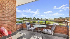 A balcony with two striped chairs and a small table, overlooking a green field and buildings. - Apollo Unit 21 - 1st Floor (Narooma)
