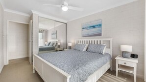 A bedroom with a bed, bedside tables, a nightstand, a ceiling fan, a mirror, and a beach-themed painting on the wall. - Apollo Unit 21 - 1st Floor (Narooma)