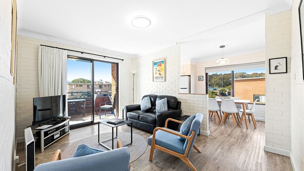 A modern living room with a black leather sofa, a television, and a dining area with white chairs. - Apollo Unit 21 - 1st Floor (Narooma)