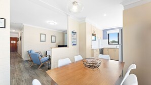 A modern kitchen with white cabinets, a wooden dining table, and a view of the outdoors. - Apollo Unit 21 - 1st Floor (Narooma)