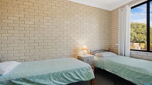 A room with two beds, a nightstand with a lamp, a brick wall, and a window with a view of trees.