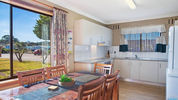 A modern kitchen with white cabinets, a dining area with wooden furniture, and a large window offering a view of the outside. - Apollo Unit 12 - Ground Floor (NAROOMA)