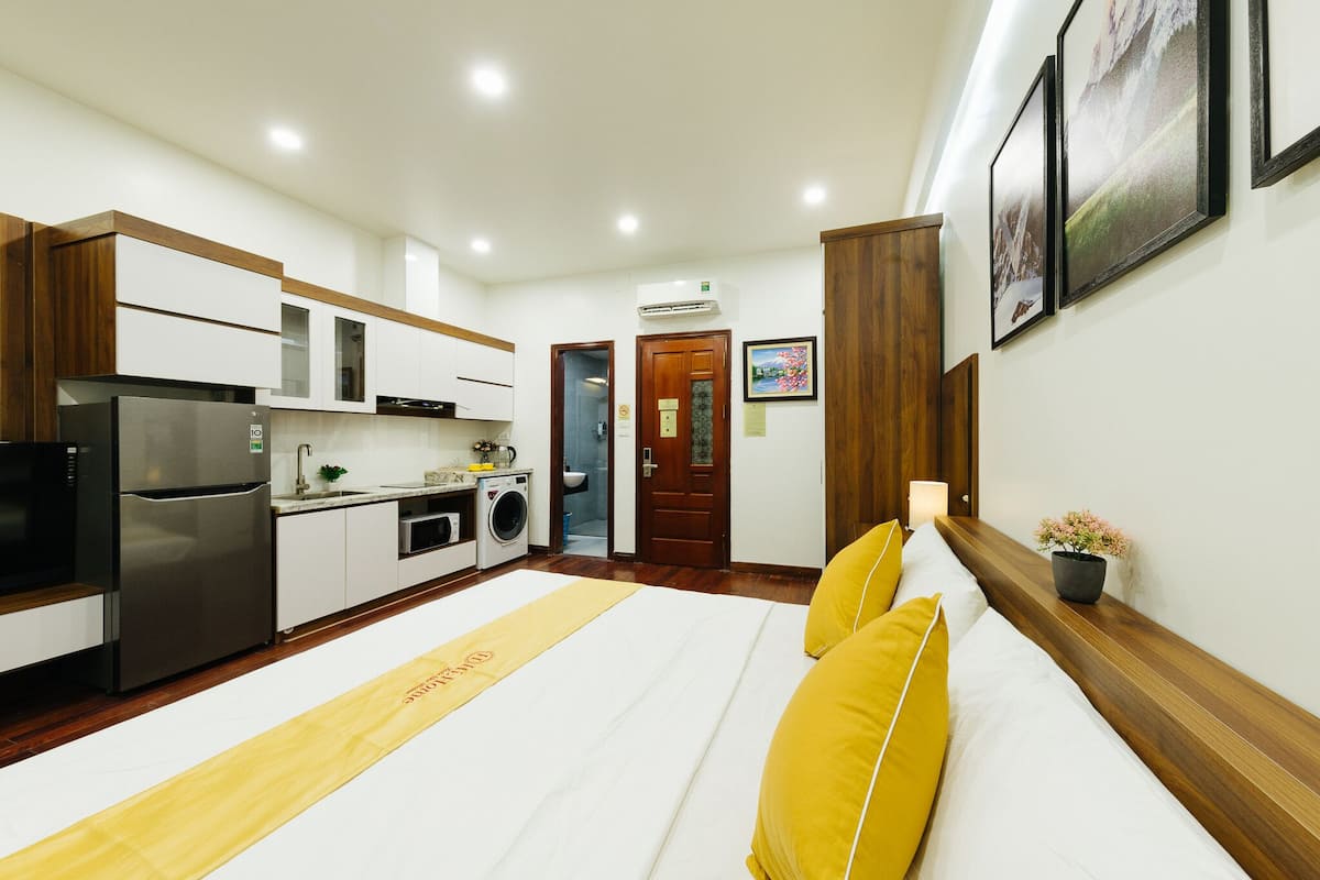 A modern hotel room with a kitchen area, a bed with yellow pillows, and a wooden bench.