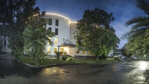 Un edificio moderno con fachada curva, rodeado de abundante vegetación y un camino de entrada mojado.