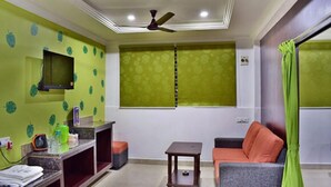 A living room with a television mounted on the wall, a ceiling fan, a sofa, a small table, and a window with patterned curtains.