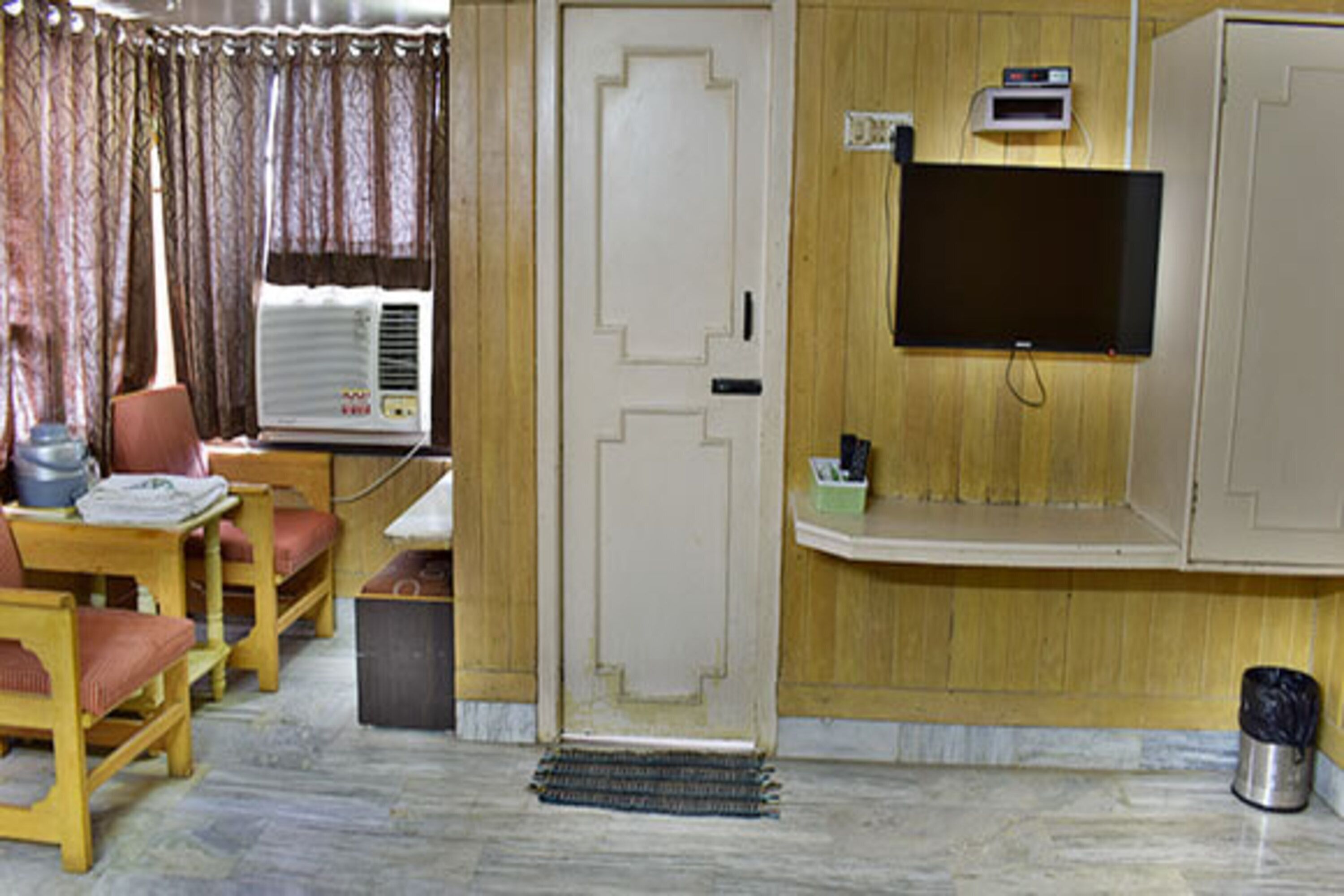 Una habitación con piso de madera, una puerta blanca, un televisor montado en la pared, una mesita con sillas y una ventana con cortinas.