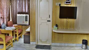 Una habitación con piso de madera, una puerta blanca, un televisor montado en la pared, una mesita con sillas y una ventana con cortinas.