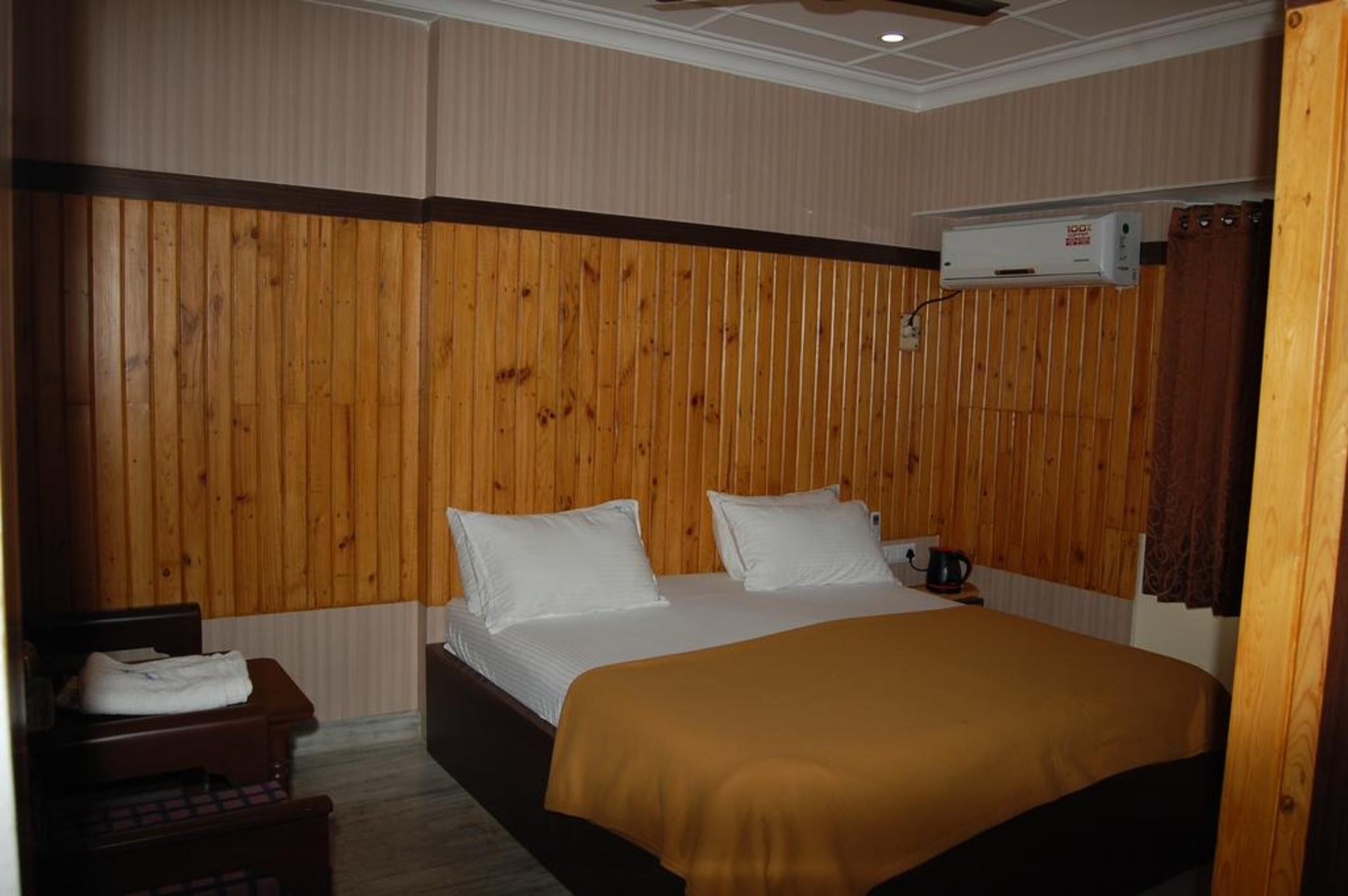 A hotel room with a wooden paneled wall, a bed with white and brown bedding, a bedside table with a lamp, and an air conditioning unit mounted on the wall.