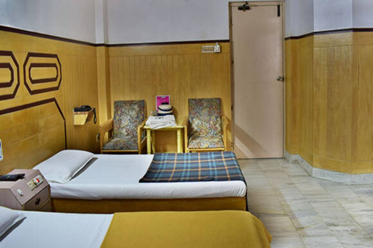 A hotel room with two beds, a small table with a lamp, two chairs, and a door.