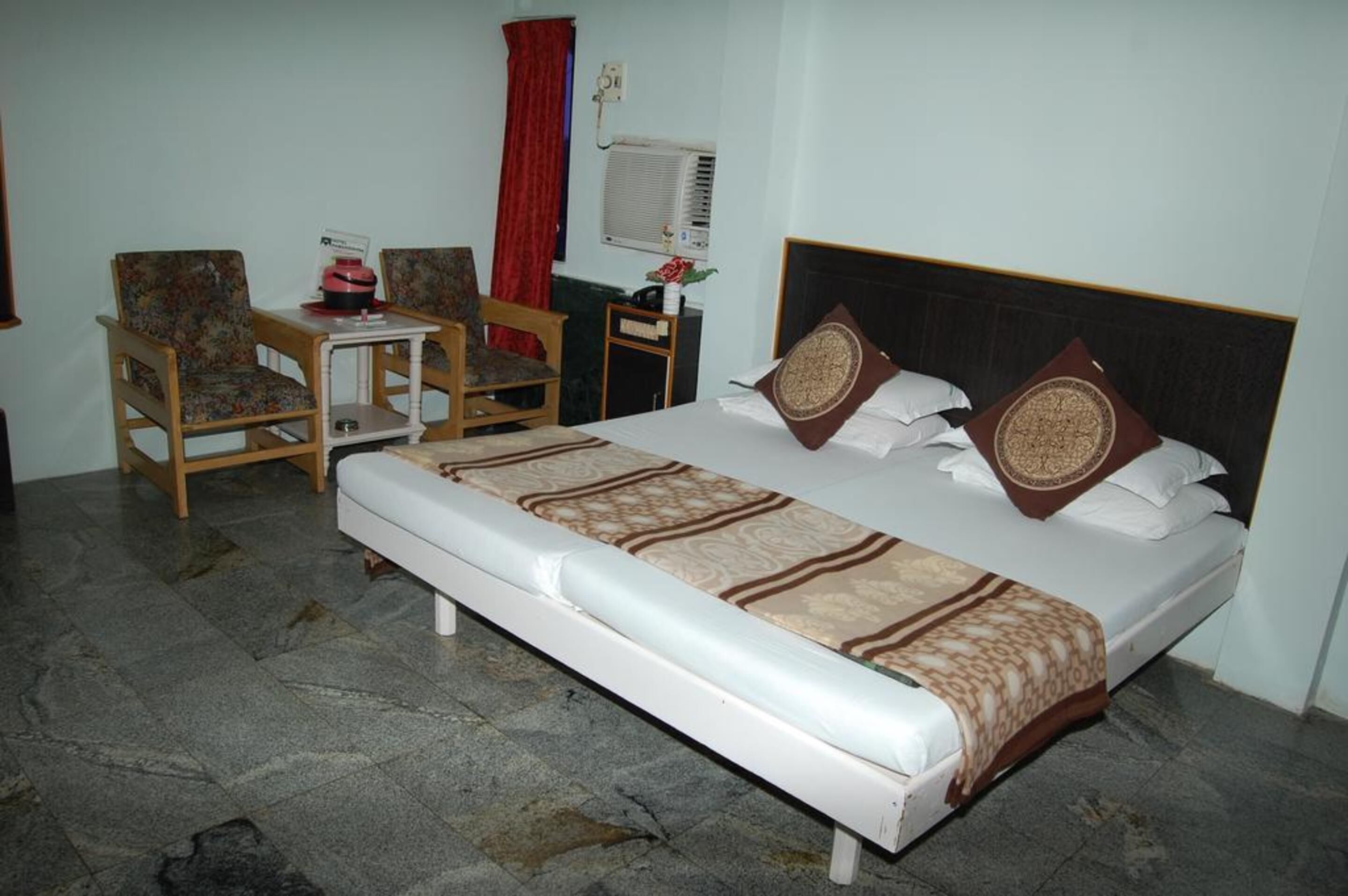 A hotel room with a single bed, two chairs, a small table, an air conditioning unit, and a red curtain.