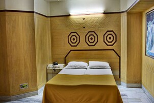 Quarto de hotel com cabeceira de madeira, cama com roupa de cama branca e parede com padrões geométricos.