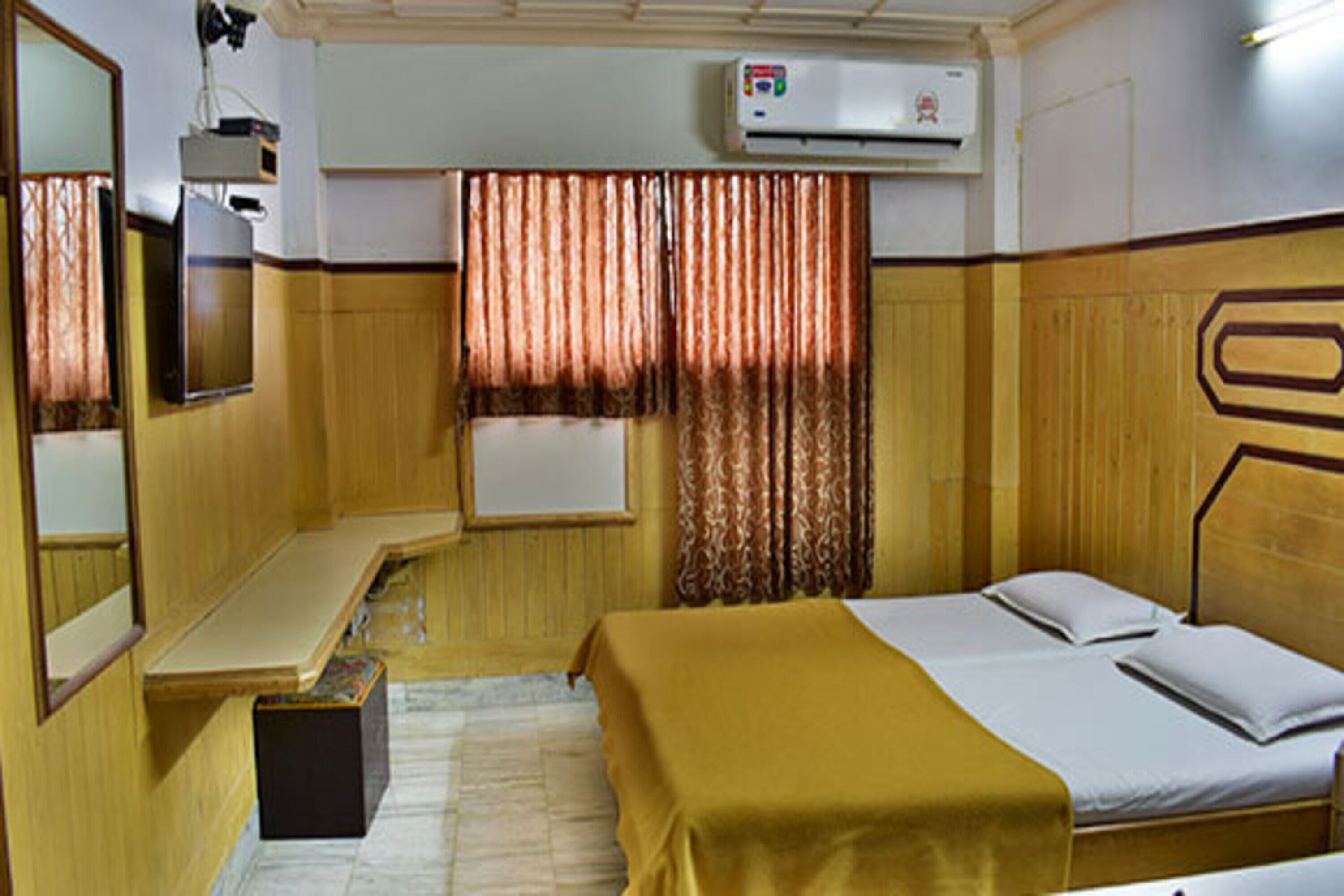 A hotel room with a bed, a bedside table, a mirror, an air conditioning unit, and a window with curtains.