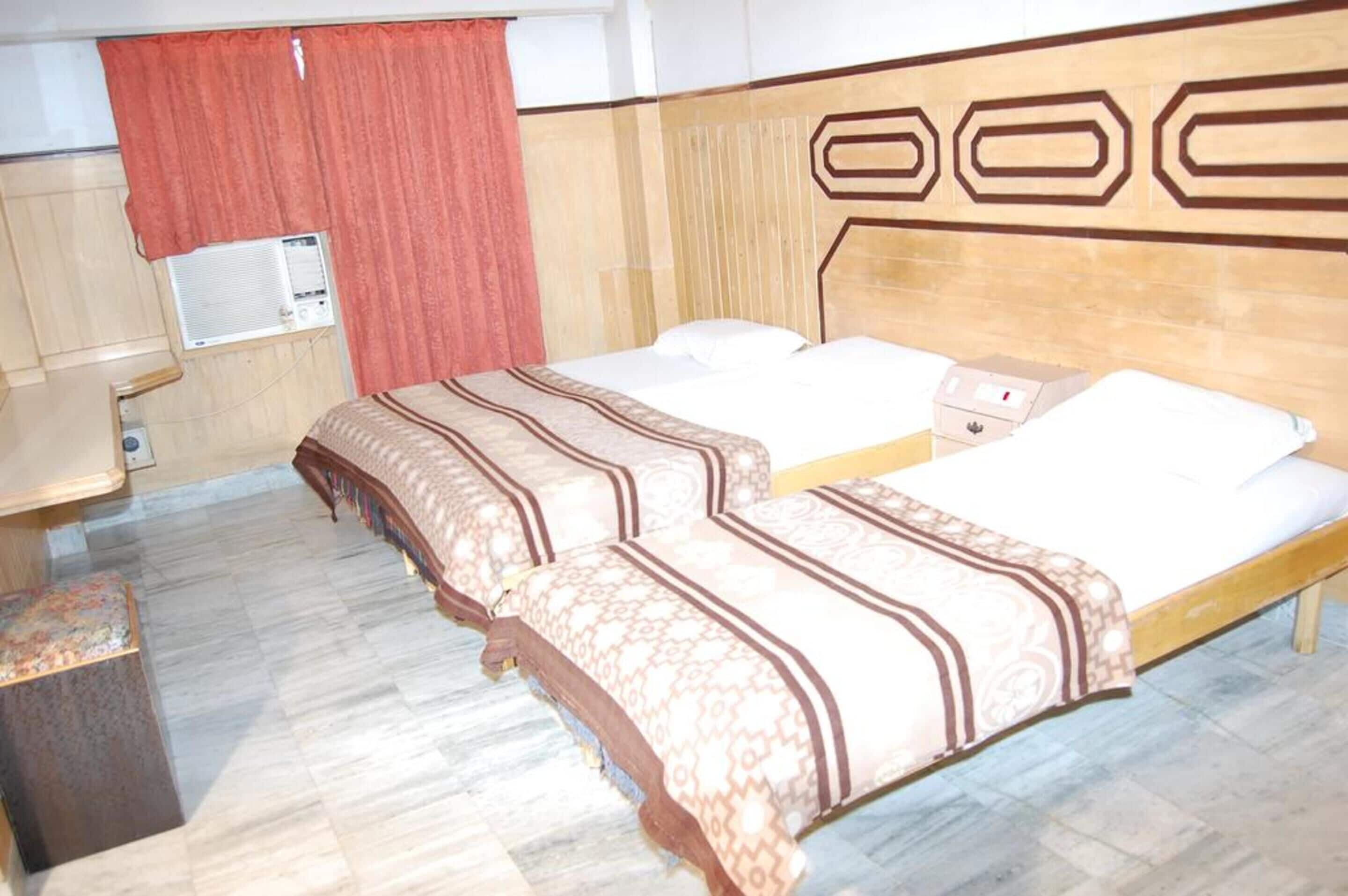 A hotel room with two beds, a wooden headboard, a small bedside table, and a red curtain.