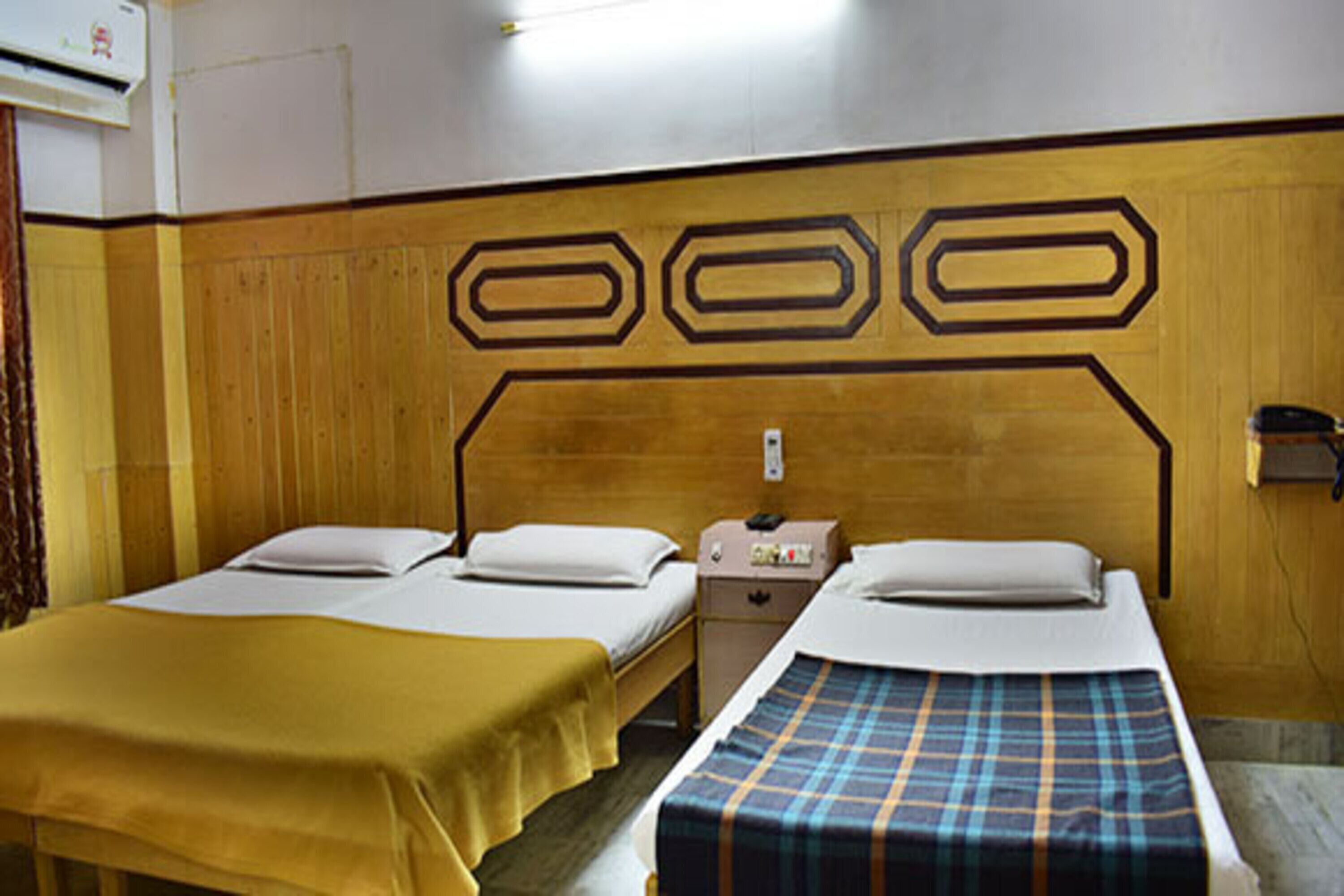 Habitación de hotel con dos camas, cabecera de madera con motivos geométricos, mesita de noche con lámpara y aire acondicionado instalado en la pared.