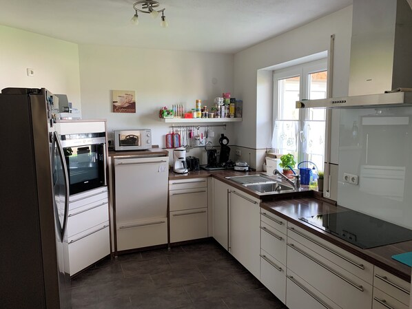 A modern kitchen with a refrigerator, oven, microwave, and a coffee maker. There are cabinets, a sink, and a window with curtains.
