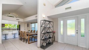 Une salle à manger spacieuse avec un plafond haut, une cuisine dotée d’une grande fenêtre et une porte blanche avec des panneaux en verre dépoli.